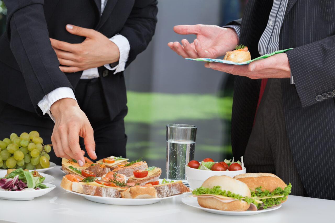 Two professional people talking, one with food in hand and other grabbing food. Sandwich, Bruschetta and water on the table