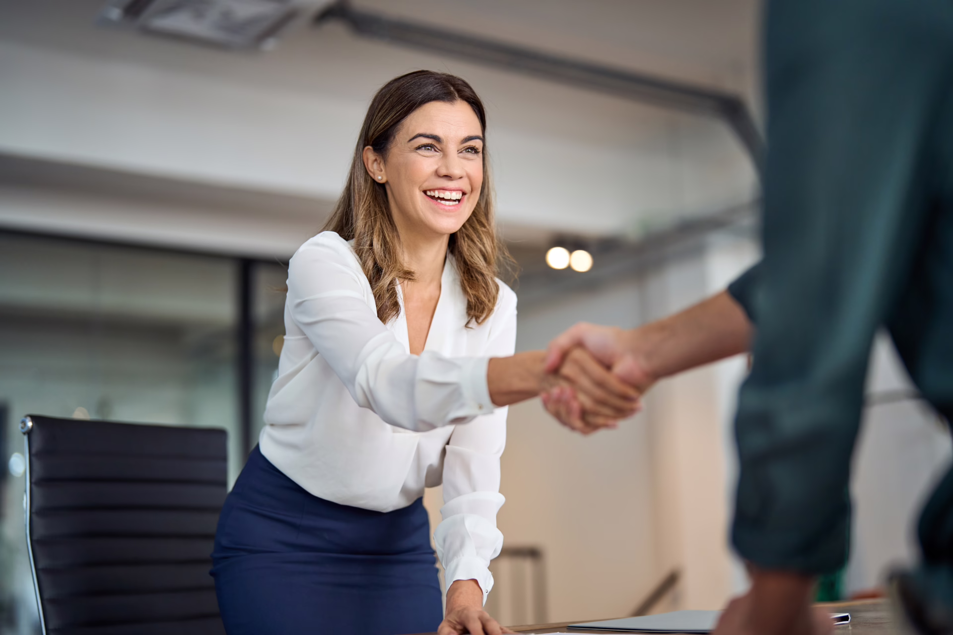 A brunette woman with yellow ends in office wear shaking a client's hand.